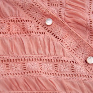 May include: Close-up of a pink cotton blouse with delicate white embroidery and a button fastening.