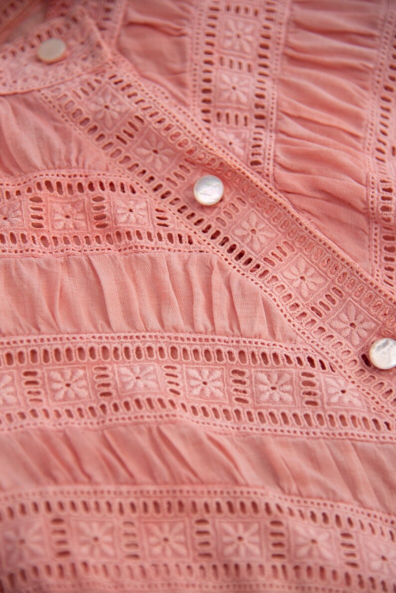 May include: Close-up of a pink cotton blouse with delicate white embroidery and a button fastening.