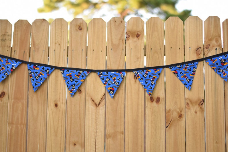May include: A blue and yellow Mickey Mouse themed banner with black trim hangs against a wooden fence.