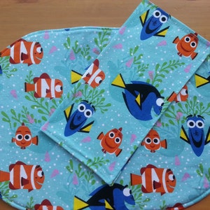 May include: A set of two blue and turquoise fabric placemats with a repeating pattern of Dory and Nemo from the movie Finding Nemo. The placemats are oval and rectangular in shape.