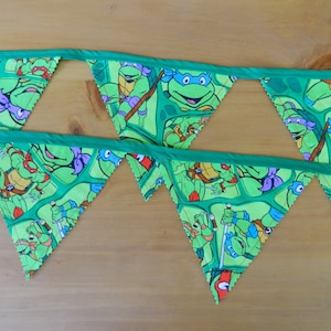 May include: Green pennant banner featuring the Teenage Mutant Ninja Turtles. Each triangular flag displays the turtles in action poses against a green background. The banner is trimmed with green fabric.