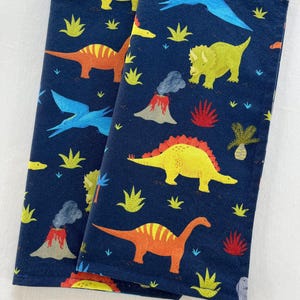 May include: Two navy blue cloth napkins with a colorful dinosaur pattern. The design includes orange, yellow, and blue dinosaurs, volcanoes, and palm trees. The napkins are folded and displayed side by side.