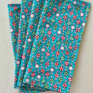 May include: Four folded teal fabric napkins with a repeating pattern of wrapped peppermint candies in red, green, and white. The candies are on a teal background. The napkins are stacked on top of each other.
