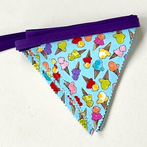 May include: A light blue fabric banner with a repeating pattern of colorful ice cream cones. The banner is trimmed with a purple border and has a purple ribbon for hanging. The ice cream cones are in various colors.