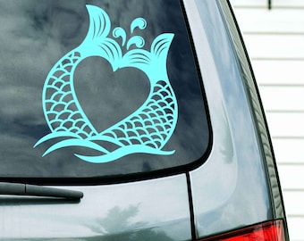 May include: A turquoise blue vinyl decal of two mermaid tails forming a heart shape with a wave design at the bottom. The decal is on the back window of a silver car.