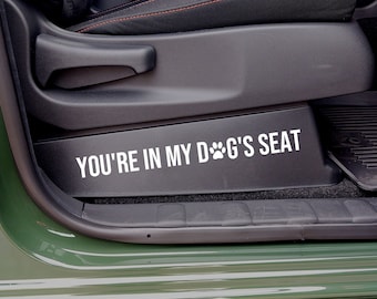 May include: A black car seat with a white sticker that says "You're in my dog's seat" with a paw print symbol.