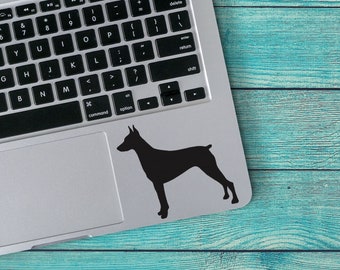 May include: A black silhouette of a Doberman Pinscher dog on a silver laptop computer.