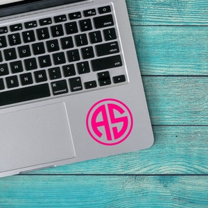 May include: A silver laptop computer with a pink monogram sticker of the letters "AS" on the top right corner of the trackpad. The laptop is on a blue wooden surface.