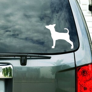Puede incluir: Una silueta blanca de un perro peque&ntilde;o, posiblemente un chihuahua, se muestra en la ventana trasera de un coche plateado. El perro est&aacute; mirando hacia la izquierda y tiene la cola enroscada.