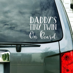 May include: A white vinyl car sticker with the text "Daddy's Tiny Twin On Board" on a silver car.