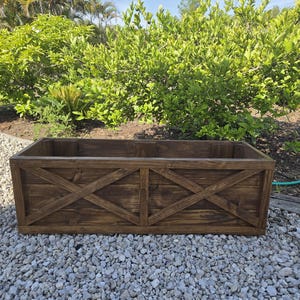 May include: A rectangular wooden planter box with a dark brown stain. The sides feature an X-shaped design. The planter is set on a bed of small, gray stones, with lush green foliage in the background.
