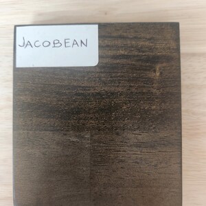 Puede incluir: Un bloque de muestra de madera cuadrado con un acabado de tinte marr&oacute;n oscuro Jacobean. Una etiqueta rectangular blanca est&aacute; adherida en la esquina superior izquierda con el texto "JACOBEAN" impreso en letras may&uacute;sculas negras, mostrando la veta.