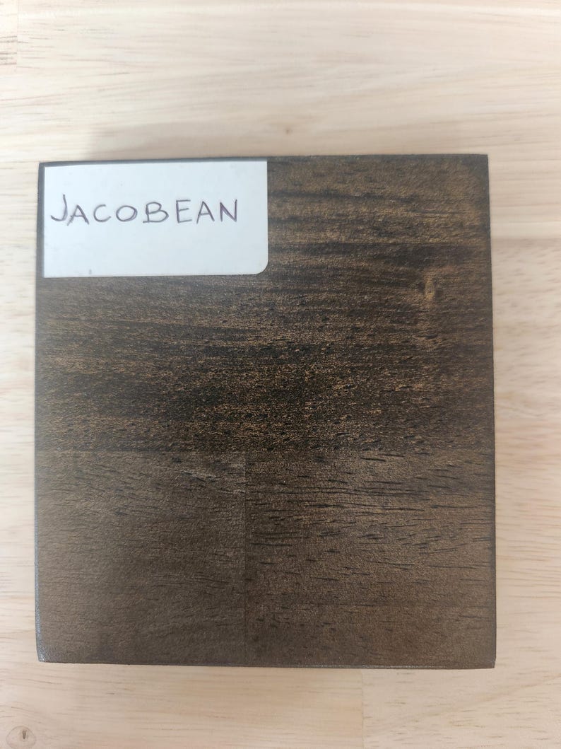 Puede incluir: Un bloque de muestra de madera cuadrado con un acabado de tinte marr&oacute;n oscuro Jacobean. Una etiqueta rectangular blanca est&aacute; adherida en la esquina superior izquierda con el texto "JACOBEAN" impreso en letras may&uacute;sculas negras, mostrando la veta.