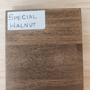 Puede incluir: Un bloque de muestra de madera cuadrado con un tinte marr&oacute;n medio y patrones de veta de madera oscura visibles. Una etiqueta rectangular blanca est&aacute; adherida en la esquina superior izquierda, mostrando claramente el texto escrito a mano "SPECIAL WALNUT".