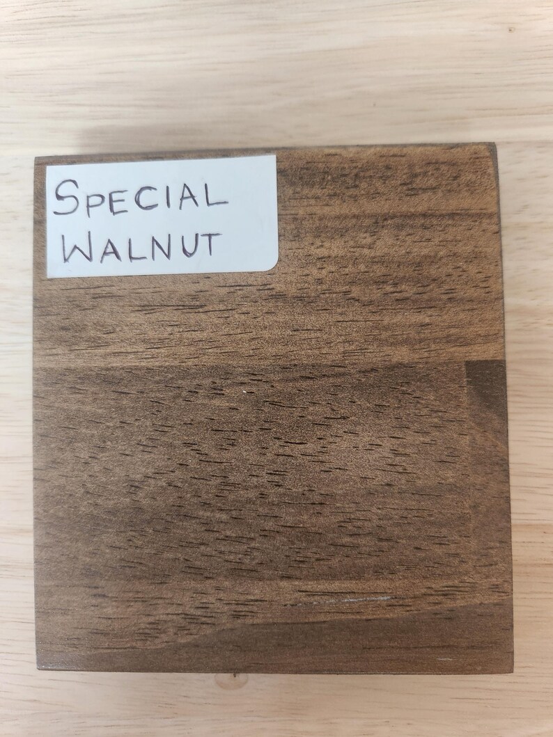 Puede incluir: Un bloque de muestra de madera cuadrado con un tinte marr&oacute;n medio y patrones de veta de madera oscura visibles. Una etiqueta rectangular blanca est&aacute; adherida en la esquina superior izquierda, mostrando claramente el texto escrito a mano "SPECIAL WALNUT".