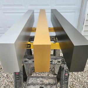 May include: Three rectangular wooden beams, painted in silver, gold, and dark gray, are displayed on a yellow and black work stand. The beams are arranged side by side, showcasing their different colors and finishes.