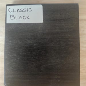 Puede incluir: Un bloque de muestra de madera cuadrado con un acabado de grano de madera negro oscuro. Una etiqueta rectangular blanca est&aacute; adherida en la esquina superior izquierda, con el texto escrito a mano "CLASSIC BLACK" en tinta negra, identificando el color.