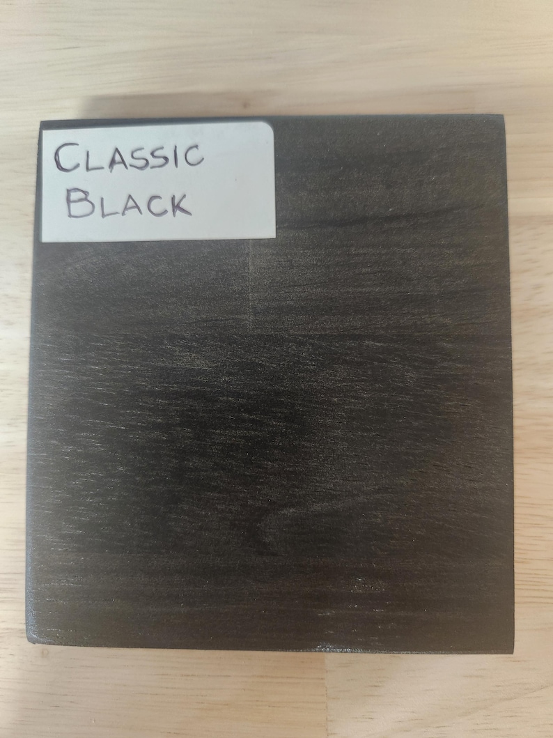 Puede incluir: Un bloque de muestra de madera cuadrado con un acabado de grano de madera negro oscuro. Una etiqueta rectangular blanca est&aacute; adherida en la esquina superior izquierda, con el texto escrito a mano "CLASSIC BLACK" en tinta negra, identificando el color.