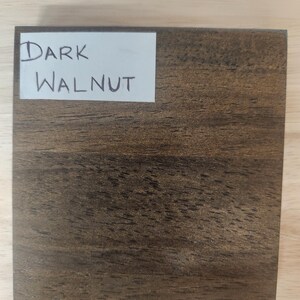 Puede incluir: Un bloque de muestra de madera cuadrado con un acabado de nogal marr&oacute;n oscuro y patrones de veta de madera natural visibles. Una etiqueta rectangular blanca est&aacute; pegada en la esquina superior izquierda con el texto Dark Walnut escrito a mano.