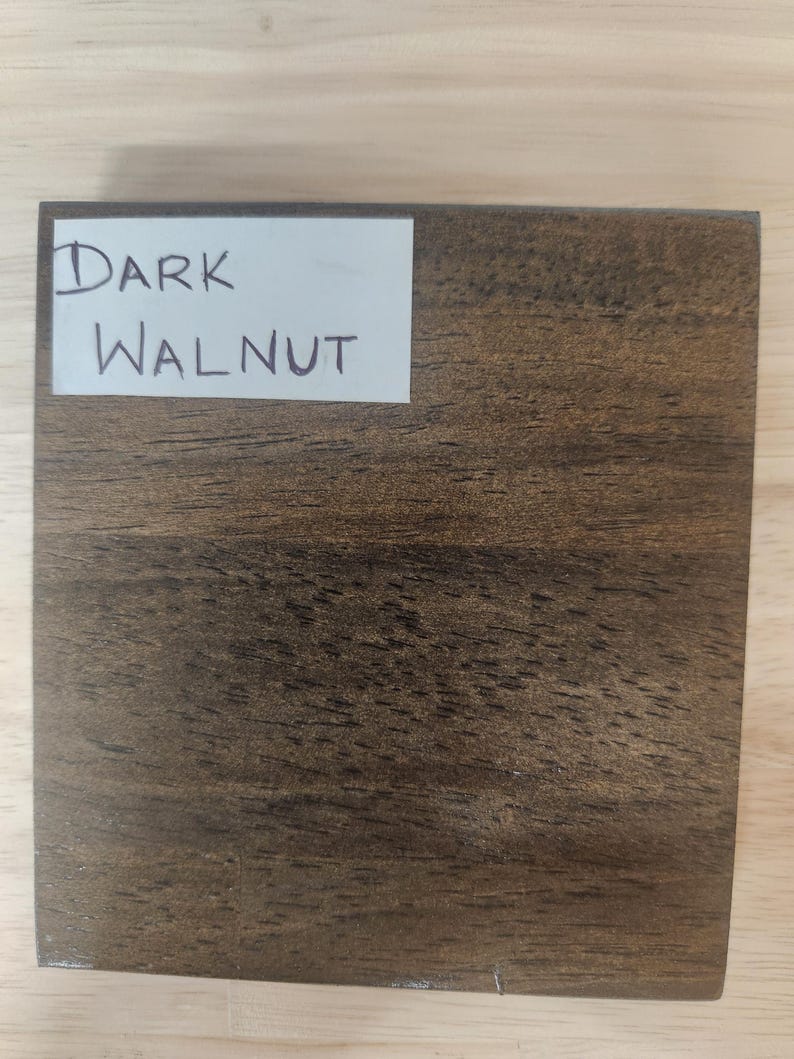 Puede incluir: Un bloque de muestra de madera cuadrado con un acabado de nogal marr&oacute;n oscuro y patrones de veta de madera natural visibles. Una etiqueta rectangular blanca est&aacute; pegada en la esquina superior izquierda con el texto Dark Walnut escrito a mano.