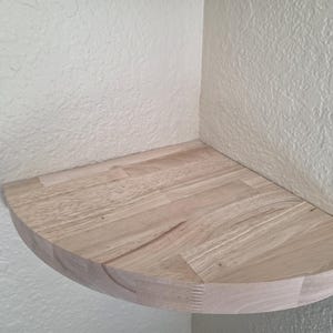 May include: A quarter-circle corner shelf made of light-colored natural wood with a visible grain pattern. The shelf features a smooth, rounded front edge and is mounted flush against two perpendicular walls, creating a flat surface for storage.