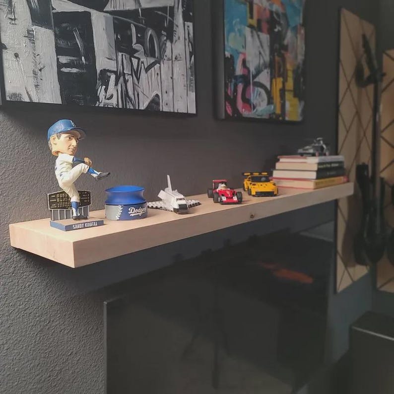 Puede incluir: Un estante flotante de madera clara que sostiene un cabez&oacute;n de Sandy Koufax, un recipiente azul de los Dodgers, un modelo de transbordador espacial blanco, un coche de carreras rojo, un coche deportivo amarillo y libros sobre una pared gris.