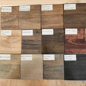 Puede incluir: Una colecci&oacute;n de doce muestras cuadradas de tinte para madera dispuestas en una cuadr&iacute;cula. Cada muestra presenta un patr&oacute;n de veta y color distintos, etiquetadas con etiquetas de papel blanco que dicen Provincial, Early American, Special Walnut, Dark Walnut, Golden Oak, Espresso, Jacobean, Red Oak, Driftwood, Weathered Oak, Classic Gray y Classic Black.