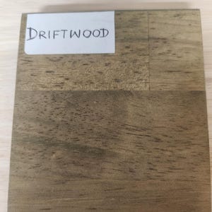 Puede incluir: Una pieza de muestra de madera cuadrada con un acabado marr&oacute;n claro desgastado y patrones de veta de madera visibles. Una etiqueta rectangular blanca est&aacute; adherida en la esquina superior izquierda con la palabra DRIFTWOOD escrita en tinta negra.