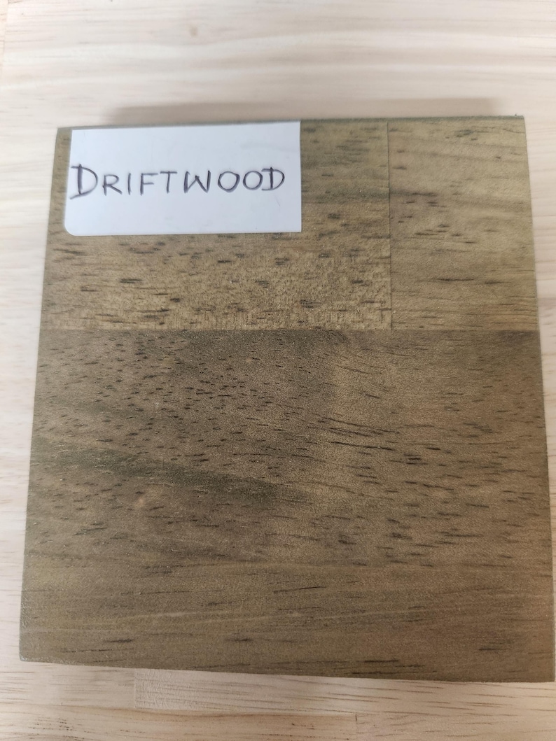 Puede incluir: Una pieza de muestra de madera cuadrada con un acabado marr&oacute;n claro desgastado y patrones de veta de madera visibles. Una etiqueta rectangular blanca est&aacute; adherida en la esquina superior izquierda con la palabra DRIFTWOOD escrita en tinta negra.