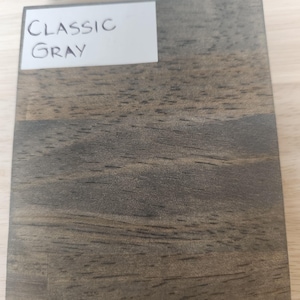 Puede incluir: Una muestra de madera rectangular con un acabado de tinte gris cl&aacute;sico, que presenta patrones de grano de madera oscura visibles y una etiqueta blanca en la esquina superior izquierda con el texto impreso en negro Classic Gray.