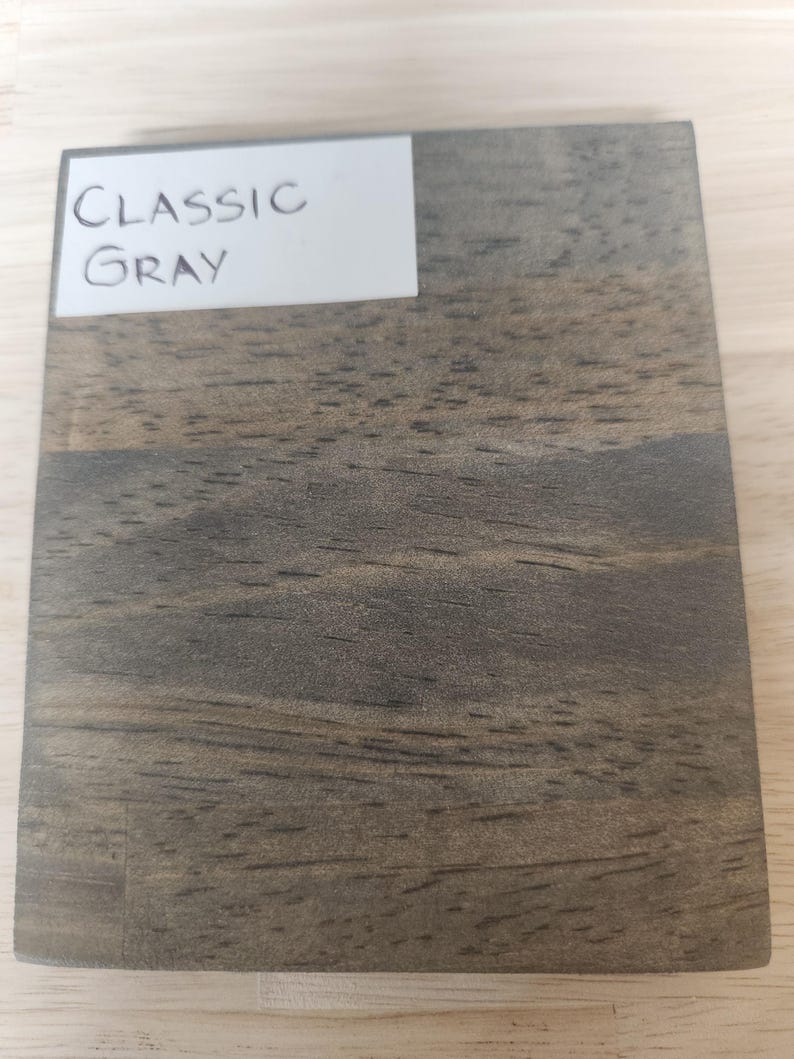 Puede incluir: Una muestra de madera rectangular con un acabado de tinte gris cl&aacute;sico, que presenta patrones de grano de madera oscura visibles y una etiqueta blanca en la esquina superior izquierda con el texto impreso en negro Classic Gray.