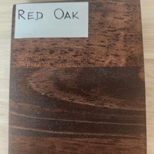 Puede incluir: Una pieza rectangular de madera te&ntilde;ida con un patr&oacute;n de veta marr&oacute;n oscuro visible. Una etiqueta rectangular blanca est&aacute; adherida en la esquina superior izquierda, con el texto escrito a mano "RED OAK" en tinta negra, identificando la madera.