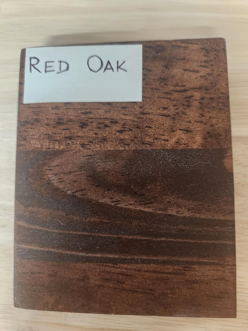 Puede incluir: Una pieza rectangular de madera te&ntilde;ida con un patr&oacute;n de veta marr&oacute;n oscuro visible. Una etiqueta rectangular blanca est&aacute; adherida en la esquina superior izquierda, con el texto escrito a mano "RED OAK" en tinta negra, identificando la madera.