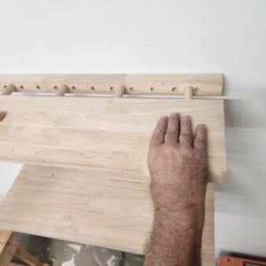 Puede incluir: Un estante de pared de madera de color claro que cuenta con una barra de montaje horizontal con m&uacute;ltiples orificios pretaladrados y tacos cil&iacute;ndricos que sostienen una tabla plana y rectangular. Se aprecia la veta de la madera natural.