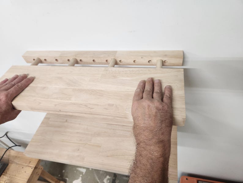Puede incluir: Un estante de pared de madera de color claro que cuenta con una barra de montaje horizontal con m&uacute;ltiples orificios pretaladrados y tacos cil&iacute;ndricos que sostienen una tabla plana y rectangular. Se aprecia la veta de la madera natural.