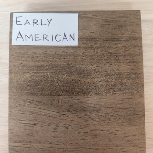 Puede incluir: Un bloque de muestra de madera cuadrado con un acabado de veta de madera marr&oacute;n medio. Una etiqueta rectangular blanca est&aacute; adherida en la esquina superior izquierda, mostrando el texto impreso en negro Early American en una fuente sans-serif.