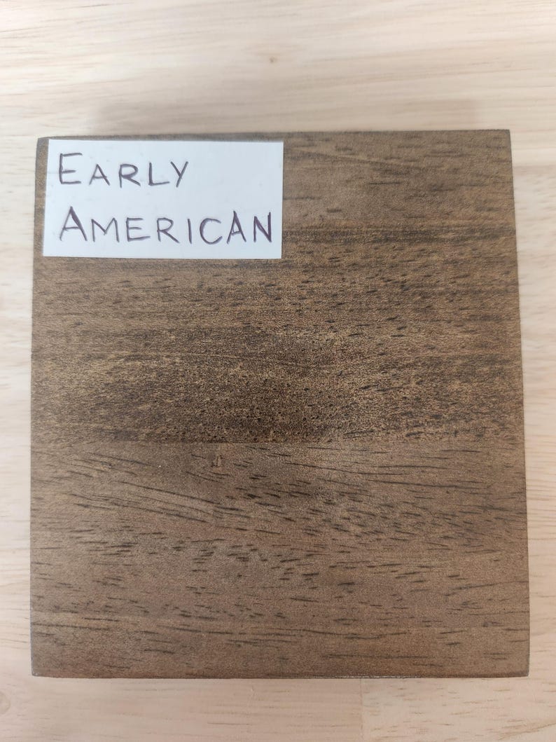 Puede incluir: Un bloque de muestra de madera cuadrado con un acabado de veta de madera marr&oacute;n medio. Una etiqueta rectangular blanca est&aacute; adherida en la esquina superior izquierda, mostrando el texto impreso en negro Early American en una fuente sans-serif.