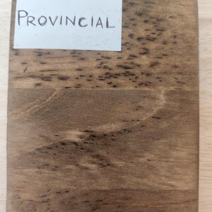 Puede incluir: Una tabla de muestra de madera rectangular con un acabado de tinte marr&oacute;n medio que presenta patrones de grano de madera oscura visibles y una etiqueta de papel blanco en la esquina superior izquierda con la palabra PROVINCIAL impresa en letras may&uacute;sculas negras.