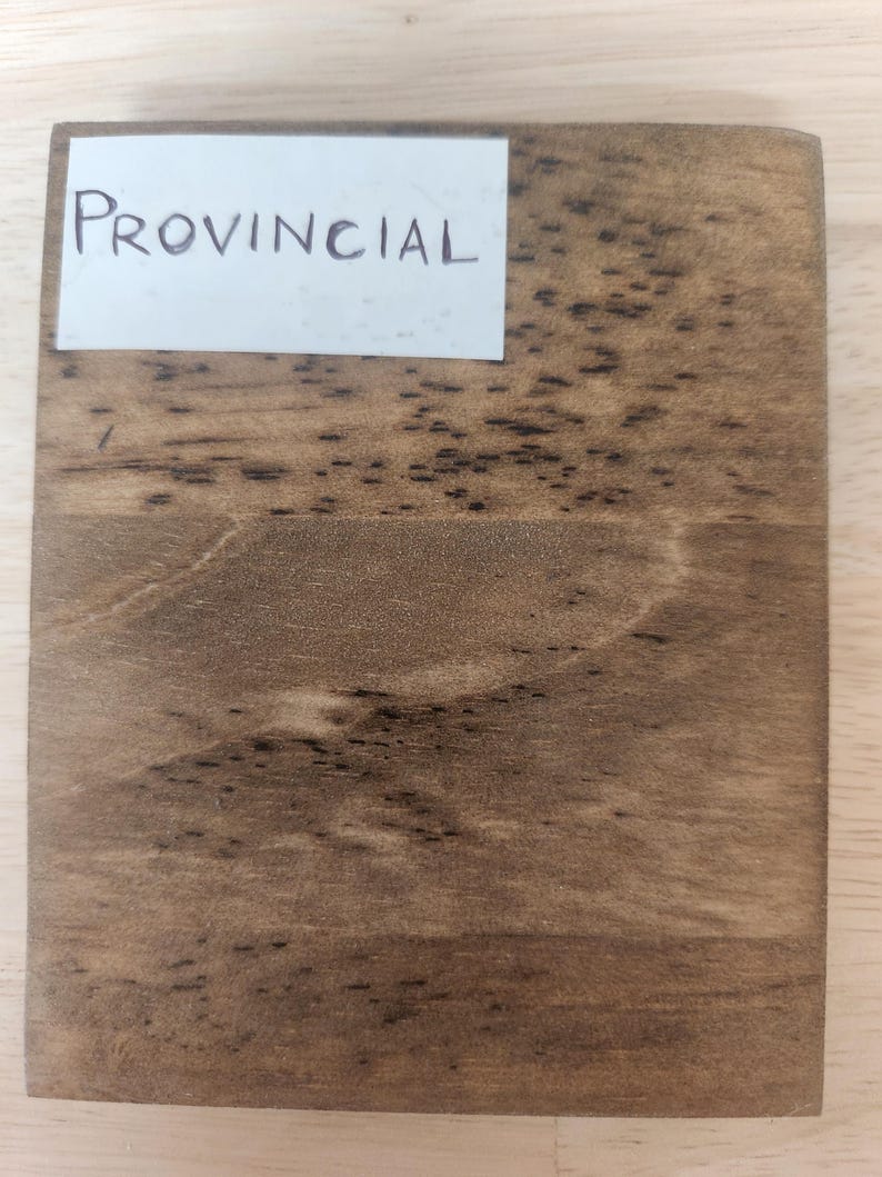 Puede incluir: Una tabla de muestra de madera rectangular con un acabado de tinte marr&oacute;n medio que presenta patrones de grano de madera oscura visibles y una etiqueta de papel blanco en la esquina superior izquierda con la palabra PROVINCIAL impresa en letras may&uacute;sculas negras.