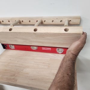 Puede incluir: Un estante de pared de madera clara con una serie de orificios circulares y tacos sobresalientes, nivelado contra una pared con una herramienta de nivel de burbuja roja Craftsman. El estante tiene un perfil rectangular con acabado de madera.