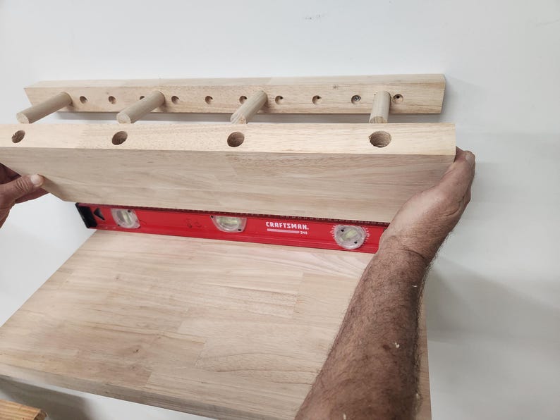 Puede incluir: Un estante de pared de madera clara con una serie de orificios circulares y tacos sobresalientes, nivelado contra una pared con una herramienta de nivel de burbuja roja Craftsman. El estante tiene un perfil rectangular con acabado de madera.