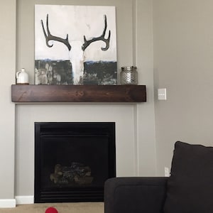 May include: A dark brown wooden mantel shelf with a white and black abstract painting of a deer skull with antlers above it. A black fireplace with a glass door is below the shelf. A black couch is in the lower right corner of the image.