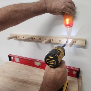 Puede incluir: Un estante de pared de madera clara con cuatro clavijas sobresalientes est&aacute; siendo instalado. Un taladro inal&aacute;mbrico amarillo y negro atornilla una fijaci&oacute;n en el estante, mientras un detector de vigas naranja se sostiene arriba. Un nivel rojo descansa debajo.