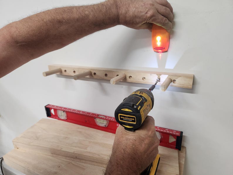 Puede incluir: Un estante de pared de madera clara con cuatro clavijas sobresalientes est&aacute; siendo instalado. Un taladro inal&aacute;mbrico amarillo y negro atornilla una fijaci&oacute;n en el estante, mientras un detector de vigas naranja se sostiene arriba. Un nivel rojo descansa debajo.