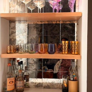 Puede incluir: Un bar dom&eacute;stico con dos estantes de madera clara sobre un salpicadero de azulejos de espejo. El estante superior contiene copas de vino transparentes e iridiscentes. El estante central muestra vasos coloridos y dorados.