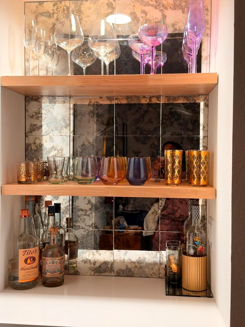 Puede incluir: Un bar dom&eacute;stico con dos estantes de madera clara sobre un salpicadero de azulejos de espejo. El estante superior contiene copas de vino transparentes e iridiscentes. El estante central muestra vasos coloridos y dorados.