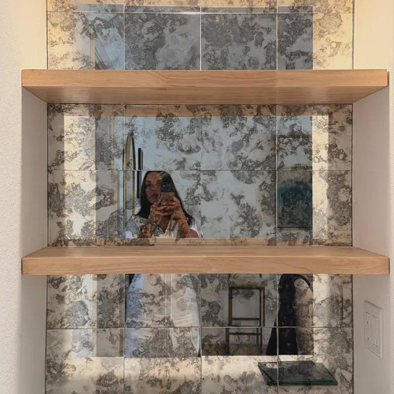 Puede incluir: Dos estantes horizontales de madera clara montados sobre una pared cubierta de azulejos de espejo cuadrados con un acabado antiguo y desgastado. La superficie espejada presenta un aspecto moteado con tonos grises y plateados.