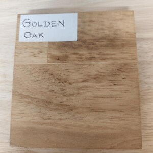Puede incluir: Un bloque de muestra de madera cuadrado que presenta un patr&oacute;n de veta de madera natural de color marr&oacute;n claro con vetas de color marr&oacute;n oscuro visibles. Una etiqueta rectangular blanca est&aacute; adherida en la esquina superior izquierda.