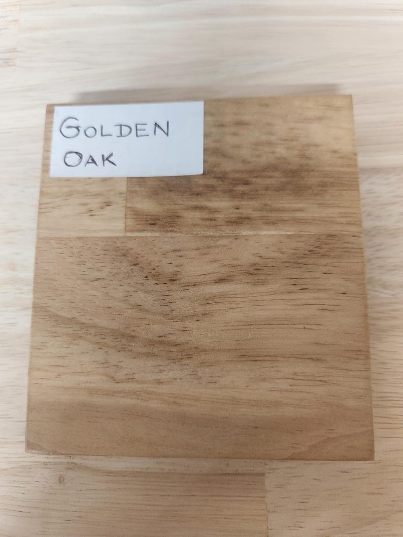 Puede incluir: Un bloque de muestra de madera cuadrado que presenta un patr&oacute;n de veta de madera natural de color marr&oacute;n claro con vetas de color marr&oacute;n oscuro visibles. Una etiqueta rectangular blanca est&aacute; adherida en la esquina superior izquierda.