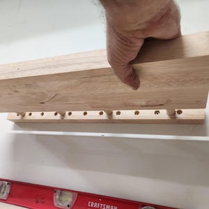 Puede incluir: Un estante de pared de madera de color claro que presenta una fila de orificios circulares espaciados uniformemente a lo largo del borde inferior, con un nivel de burbuja rojo marca Craftsman colocado debajo para asegurar la alineaci&oacute;n.